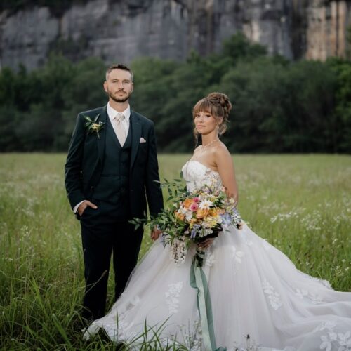 Bride in a white lace gown and groom in a dark green suit walk hand in hand across a grassy field, with trees behind them.