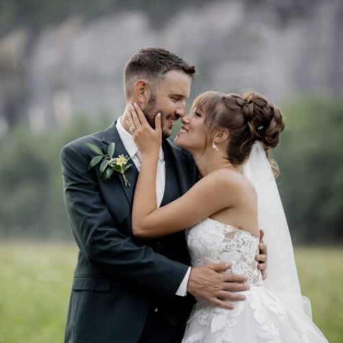 Bride and groom share a kiss outdoors, the bride in a lace wedding gown and veil, the groom in a dark suit with a boutonniere.