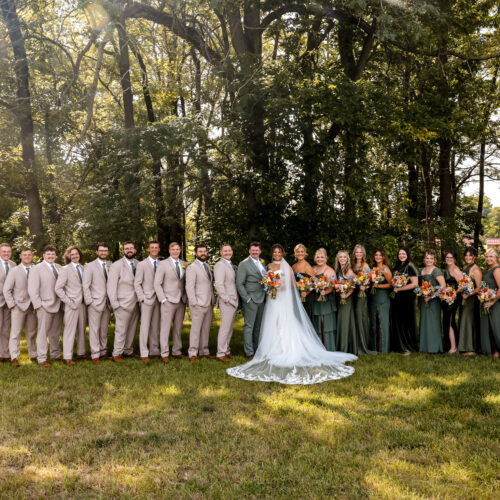 Bride and groom at center of a long wedding party lineup outdoors among tall trees in sunlight.