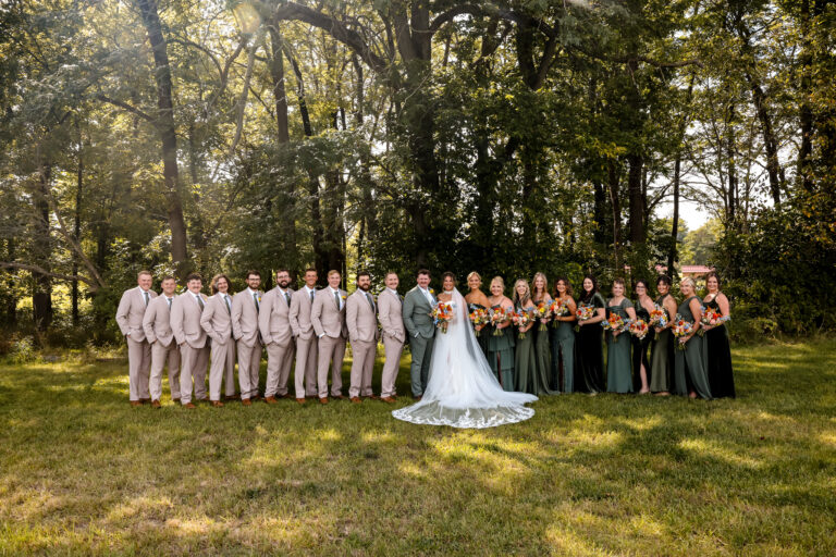 Bride and groom at center of a long wedding party lineup outdoors among tall trees in sunlight.