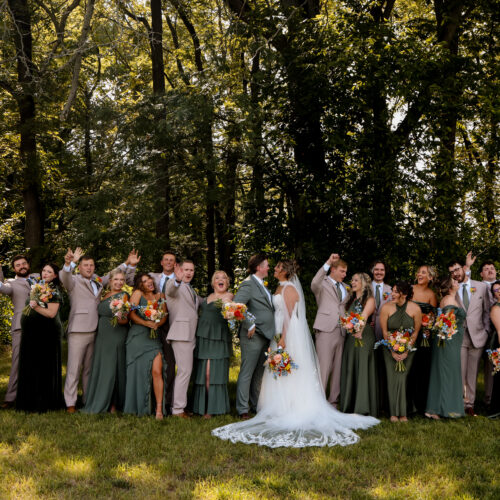 Bride and groom center with a large wedding party outdoors, bridesmaids in green gowns and groomsmen in light suits, holding bouquets amid trees.