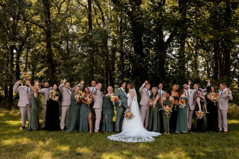 Bride and groom center with a large wedding party outdoors, bridesmaids in green gowns and groomsmen in light suits, holding bouquets amid trees.