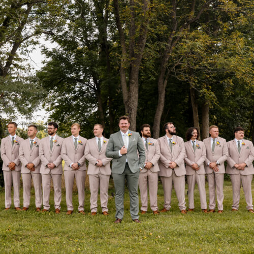 Groom in a green suit standing with his groomsmen in beige suits outdoors among trees at a wedding photo line.