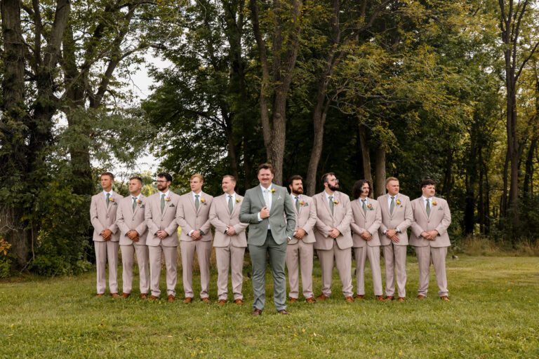 Groom in a green suit standing with his groomsmen in beige suits outdoors among trees at a wedding photo line.