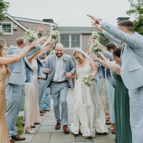 Bride and groom walk down a stone path while bridesmaids and groomsmen in pastel blue outfits raise bouquets in celebration outside a house.
