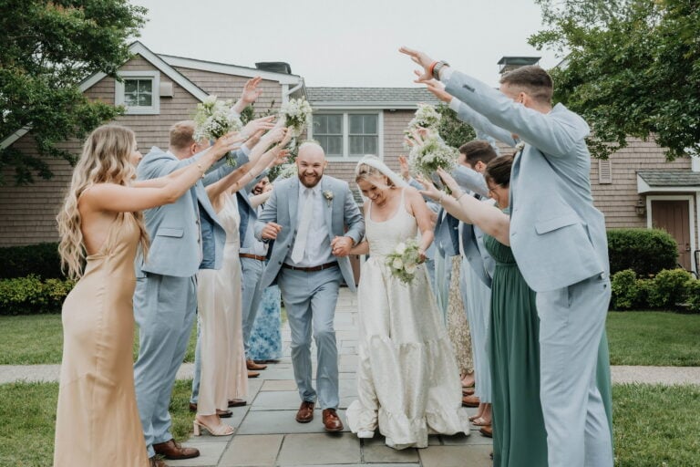 Bride and groom walk down a stone path while bridesmaids and groomsmen in pastel blue outfits raise bouquets in celebration outside a house.