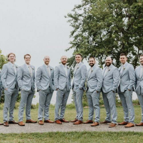 Group of groomsmen in light-blue suits posing outdoors on a green lawn, brown shoes, with trees in the background.
