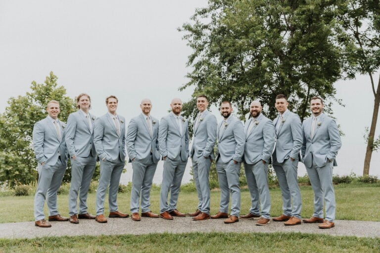 Group of groomsmen in light-blue suits posing outdoors on a green lawn, brown shoes, with trees in the background.