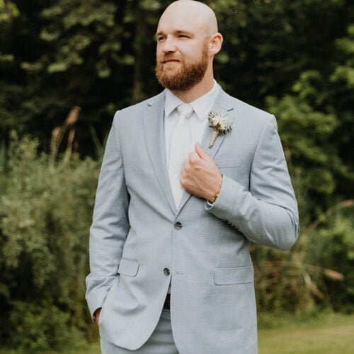 Man with a trimmed beard and bald head wearing a light gray suit, white shirt, and tie, standing outdoors with greenery in the background and a boutonniere on his lapel.",
