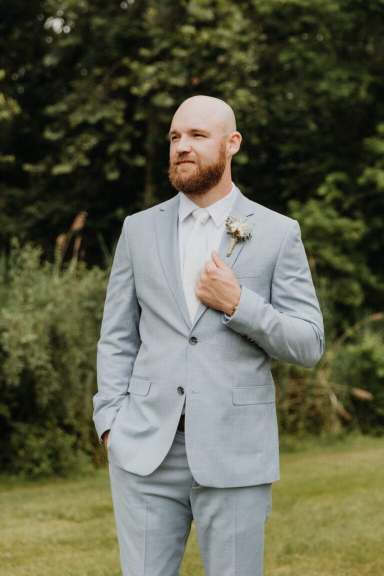 Man with a trimmed beard and bald head wearing a light gray suit, white shirt, and tie, standing outdoors with greenery in the background and a boutonniere on his lapel.",