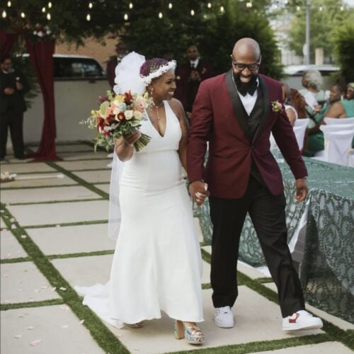 Bride in a white gown holding a bouquet walks hand in hand with a smiling groom in a burgundy jacket at an outdoor wedding ceremony. 2. Smiling bride with bouquet and groom in a burgundy suit walk down the aisle at an outdoor ceremony. 3. Couple exits the ceremony together: bride with white dress and floral bouquet, groom in a maroon jacket. 4. Outdoor wedding scene: bride and groom walking along a decorated aisle with guests in the background. 5. Bride and groom holding hands as they walk through a garden ceremony space.