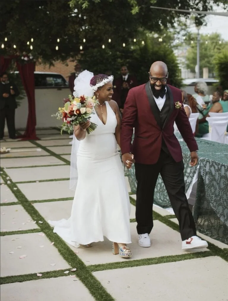 Bride in a white gown holding a bouquet walks hand in hand with a smiling groom in a burgundy jacket at an outdoor wedding ceremony. 2. Smiling bride with bouquet and groom in a burgundy suit walk down the aisle at an outdoor ceremony. 3. Couple exits the ceremony together: bride with white dress and floral bouquet, groom in a maroon jacket. 4. Outdoor wedding scene: bride and groom walking along a decorated aisle with guests in the background. 5. Bride and groom holding hands as they walk through a garden ceremony space.