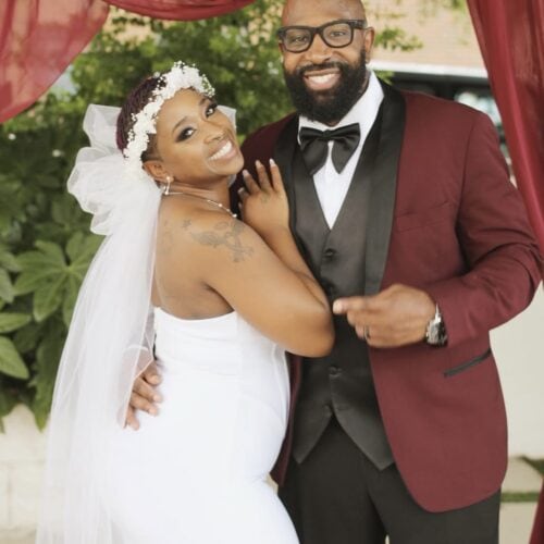 Bride and groom smiling together under burgundy drapes, man in burgundy suit jacket and black bow tie, woman in white wedding dress with veil.