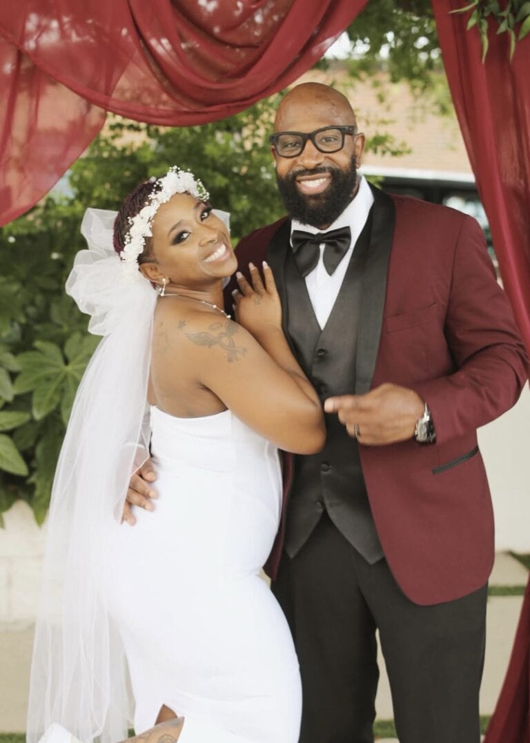 Bride and groom smiling together under burgundy drapes, man in burgundy suit jacket and black bow tie, woman in white wedding dress with veil.