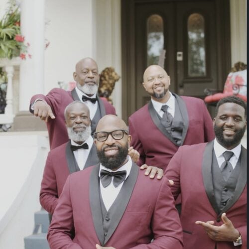 Five men in burgundy tuxedos and black bow ties pose on the front steps of a house, smiling at the camera.