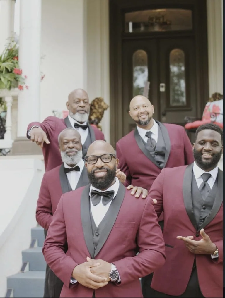 Five men in burgundy tuxedos and black bow ties pose on the front steps of a house, smiling at the camera.