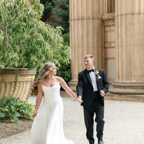 Bride in a white wedding gown and groom in a black tuxedo walk hand in hand beside stone columns amidst greenery outside the venue.