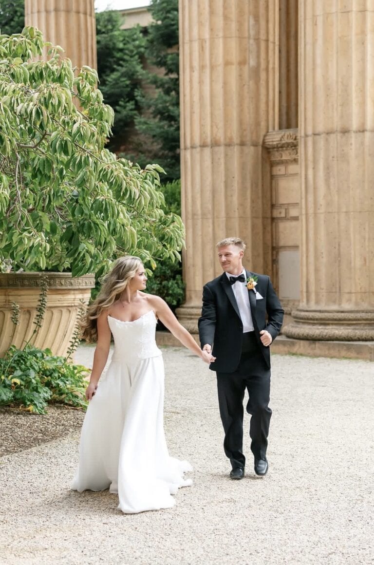 Bride in a white wedding gown and groom in a black tuxedo walk hand in hand beside stone columns amidst greenery outside the venue.