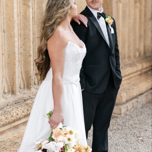 Bride in a white strapless wedding gown stands beside a groom in a black tuxedo, both looking at each other, holding a peach-and-white bouquet.