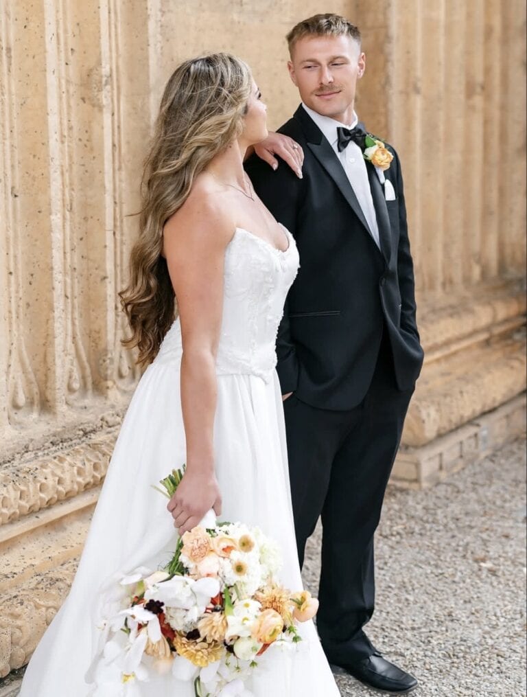Bride in a white strapless wedding gown stands beside a groom in a black tuxedo, both looking at each other, holding a peach-and-white bouquet.