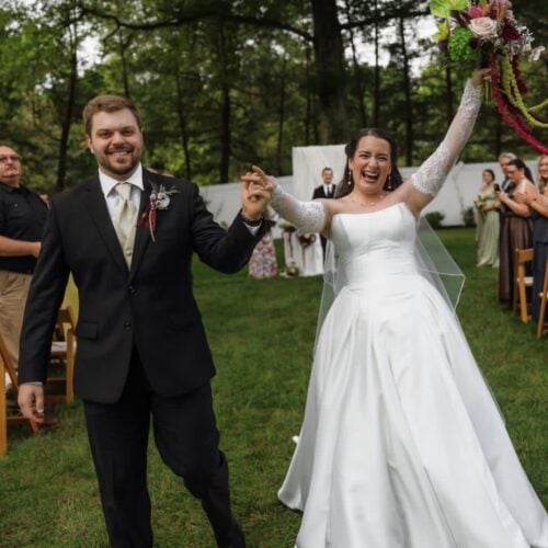 Bride and groom walk hand in hand down the aisle outdoors, the bride lifting a bouquet and smiling broadly amid guests and greenery.