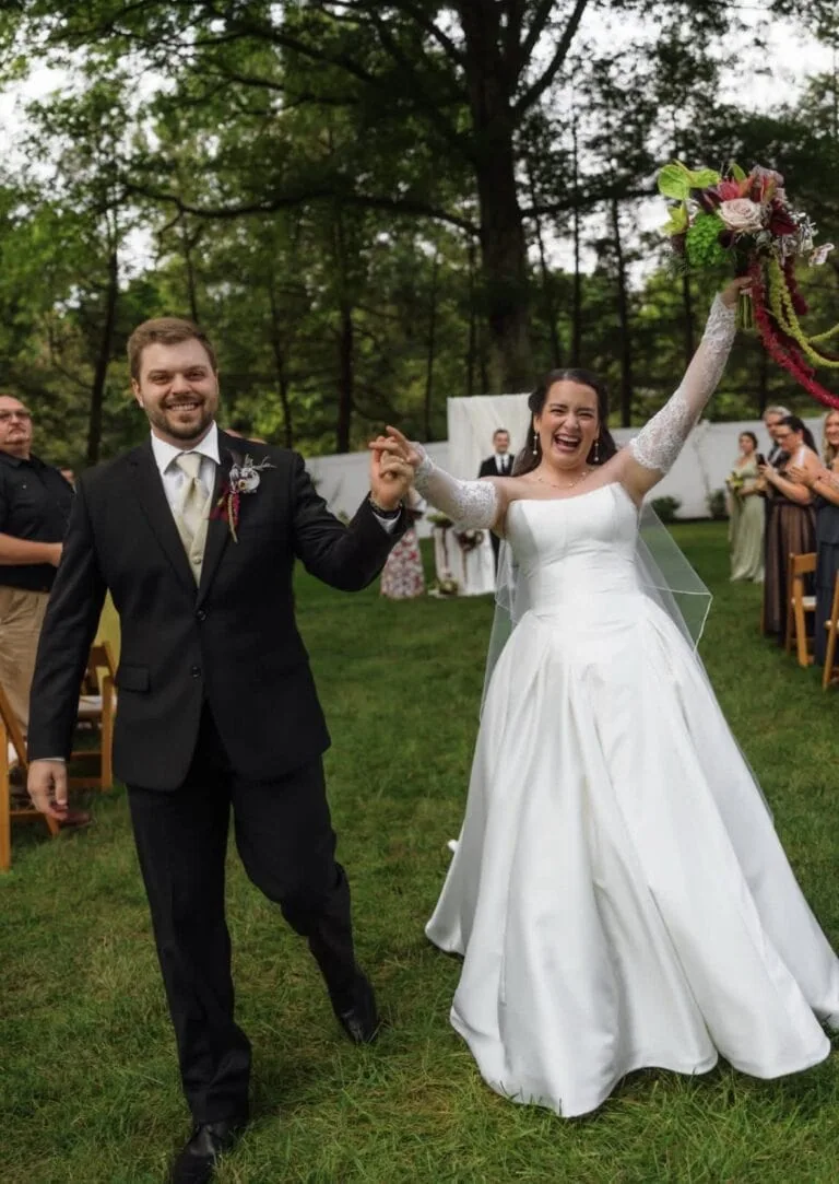 Screenshot | National Tuxedo Rentals Bride and groom walk hand in hand down the aisle outdoors, the bride lifting a bouquet and smiling broadly amid guests and greenery.