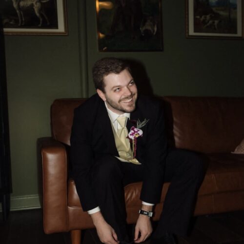 Smiling man in a black suit with a cream tie and orchid boutonniere sits on a brown leather sofa indoors, tying his shoes.