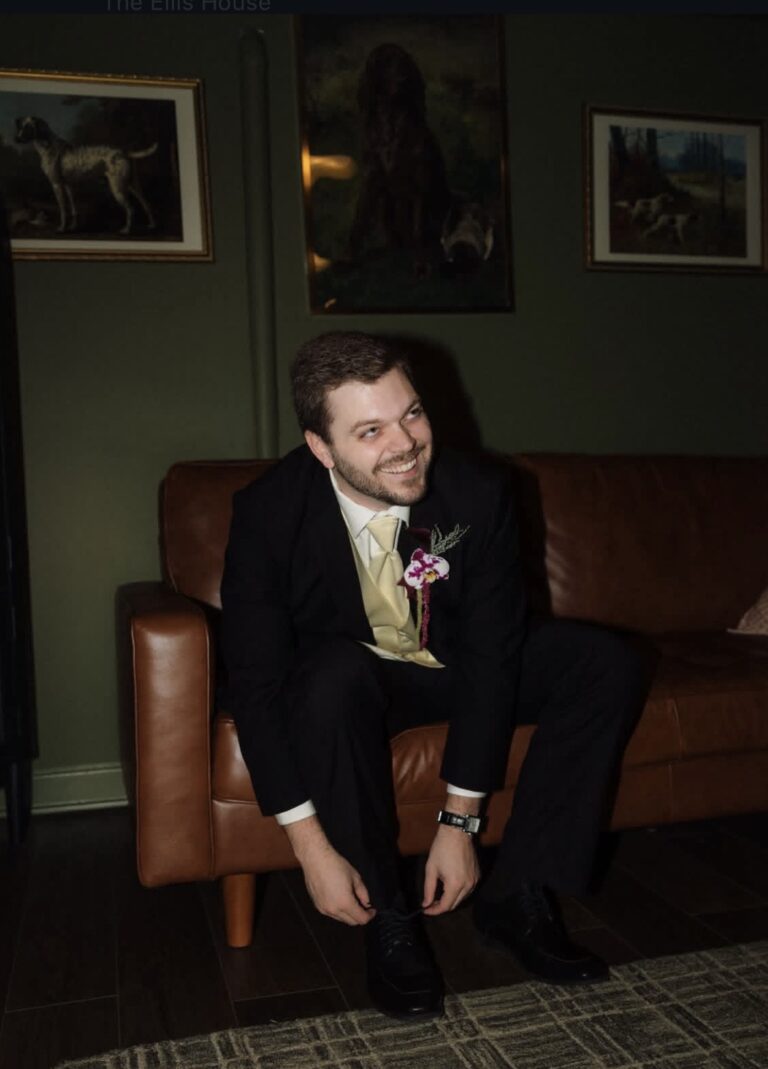 Screenshot | National Tuxedo Rentals Smiling man in a black suit with a cream tie and orchid boutonniere sits on a brown leather sofa indoors, tying his shoes.