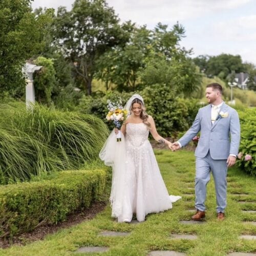 Bride in a white wedding gown holding a bouquet and veil, walking hand in hand with a groom in a light blue suit along a garden path.
