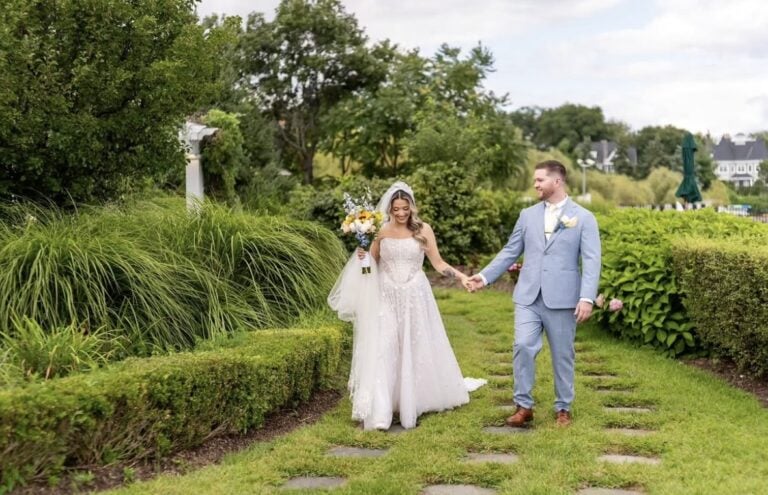 Bride in a white wedding gown holding a bouquet and veil, walking hand in hand with a groom in a light blue suit along a garden path.
