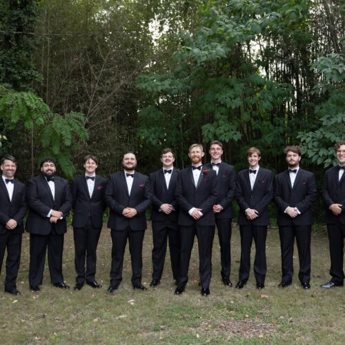 Group of men in black tuxedos and bow ties standing in a line outdoors in a leafy setting.