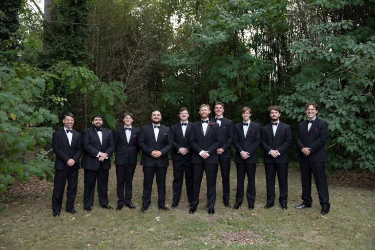 The Hines | National Tuxedo Rentals Group of men in black tuxedos and bow ties standing in a line outdoors in a leafy setting.