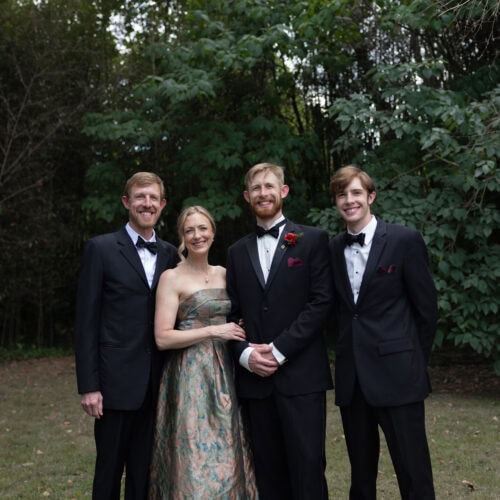 Four adults in formal attire posing outdoors in front of leafy trees.