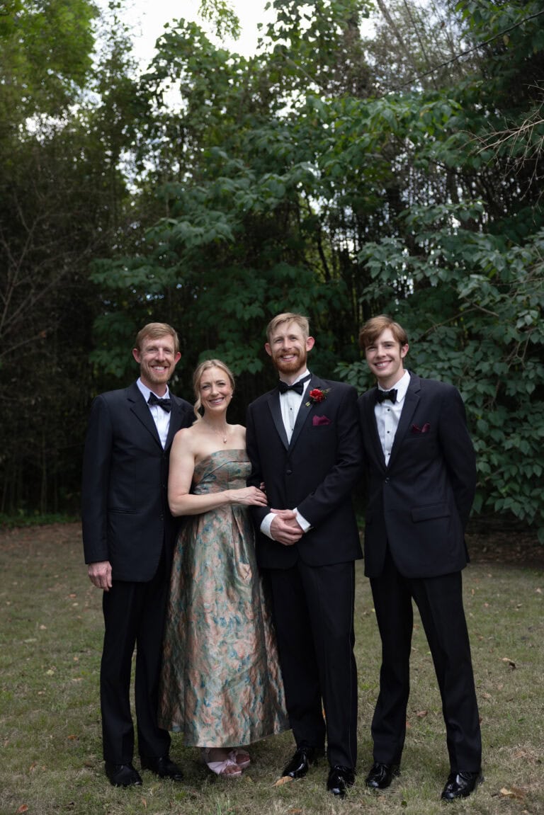 The Hines | National Tuxedo Rentals Four adults in formal attire posing outdoors in front of leafy trees.