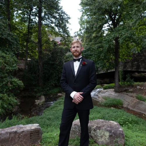 Man in a black tuxedo and bow tie stands on a stone path outdoors, with trees and greenery surrounding him and a red boutonniere at the lapel.