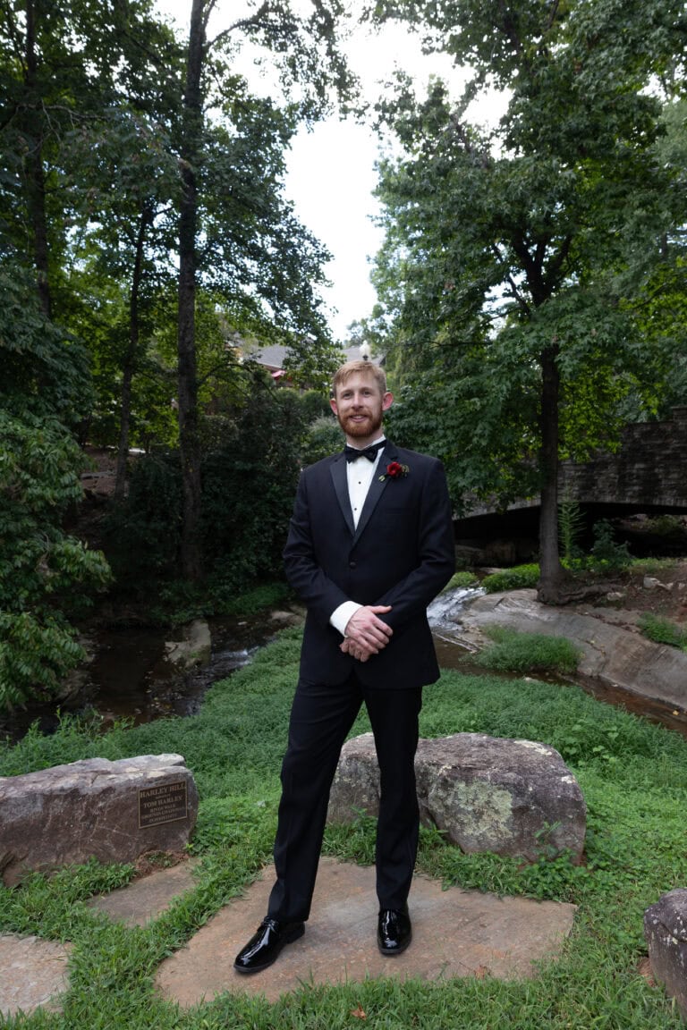 The Hines | National Tuxedo Rentals Man in a black tuxedo and bow tie stands on a stone path outdoors, with trees and greenery surrounding him and a red boutonniere at the lapel.