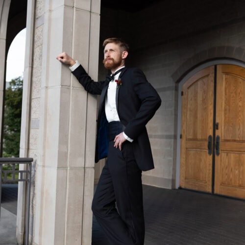 Bearded man in a black tuxedo and bow tie leaning against a stone column outside a building with a wooden door behind him.