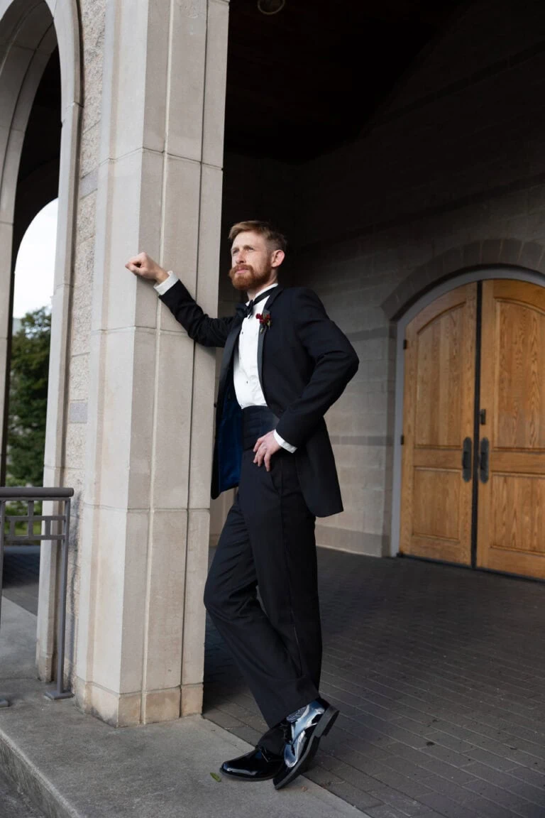 The Hines | National Tuxedo Rentals Bearded man in a black tuxedo and bow tie leaning against a stone column outside a building with a wooden door behind him.
