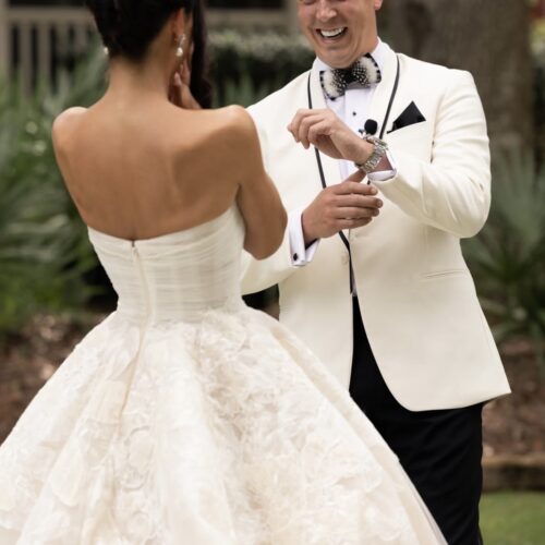 Bride and groom dancing outdoors; bride wears a strapless lace wedding gown, and the groom wears a white tux and bow tie, smiling. (Back view)