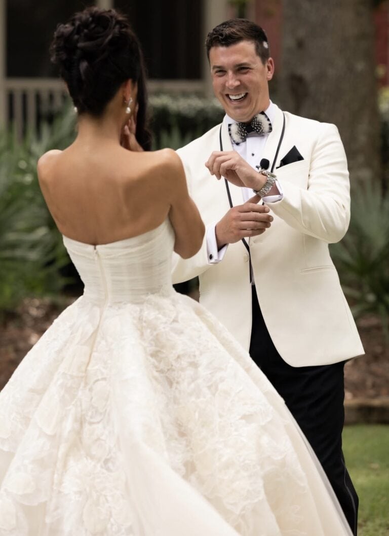 Screenshot | National Tuxedo Rentals Bride and groom dancing outdoors; bride wears a strapless lace wedding gown, and the groom wears a white tux and bow tie, smiling. (Back view)