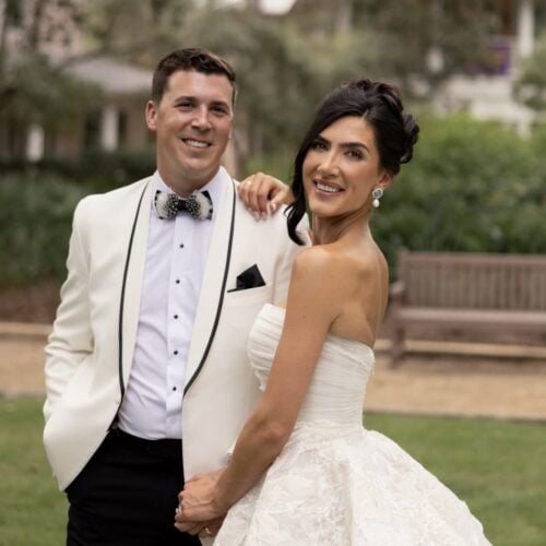 Smiling bride in a lace white wedding gown stands beside a groom in a white tuxedo with a black bow tie outdoors.
