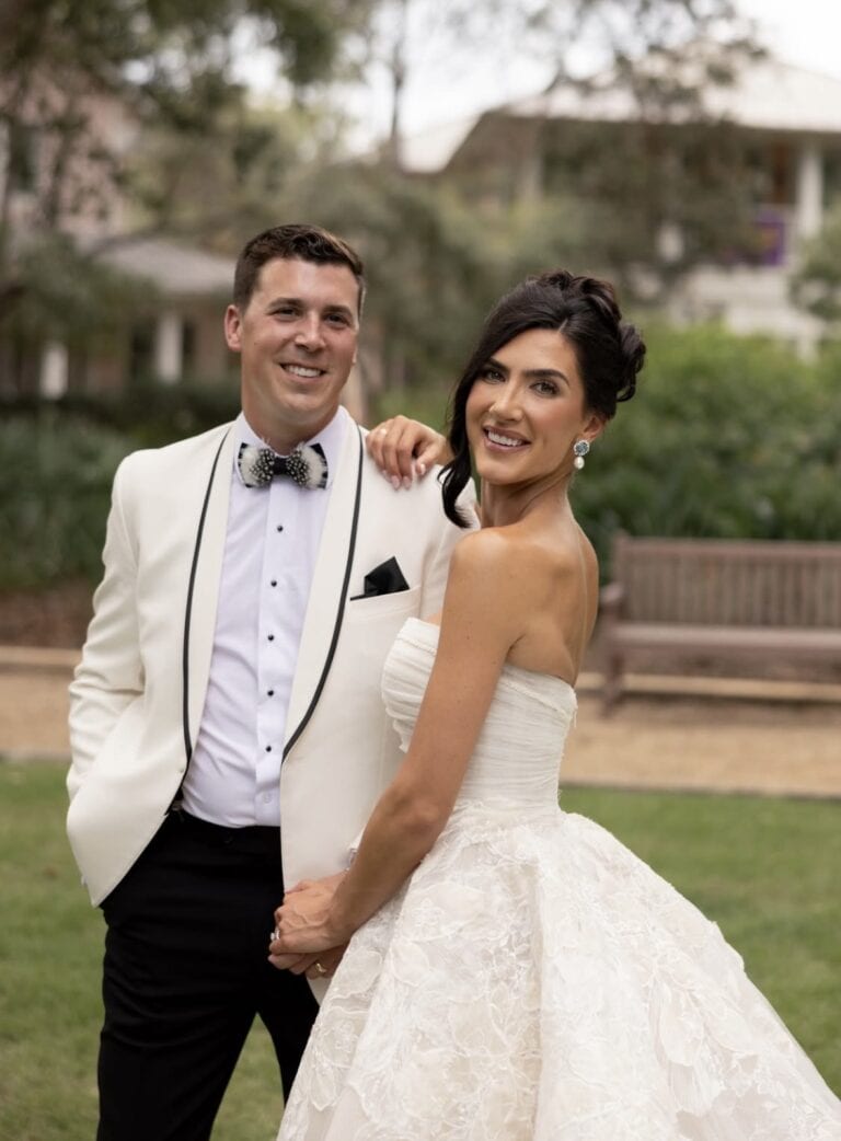 Screenshot | National Tuxedo Rentals Smiling bride in a lace white wedding gown stands beside a groom in a white tuxedo with a black bow tie outdoors.