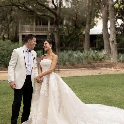 Bride in a strapless wedding gown and groom in a cream tuxedo hold hands and smile on a grassy lawn.