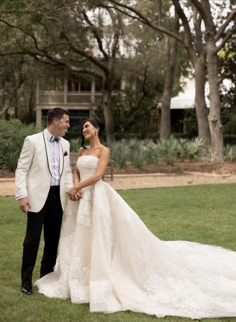 Screenshot | National Tuxedo Rentals Bride in a strapless wedding gown and groom in a cream tuxedo hold hands and smile on a grassy lawn.