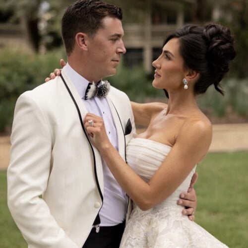 Bride in a lace wedding gown and groom in a white tuxedo embrace outdoors at a garden wedding.