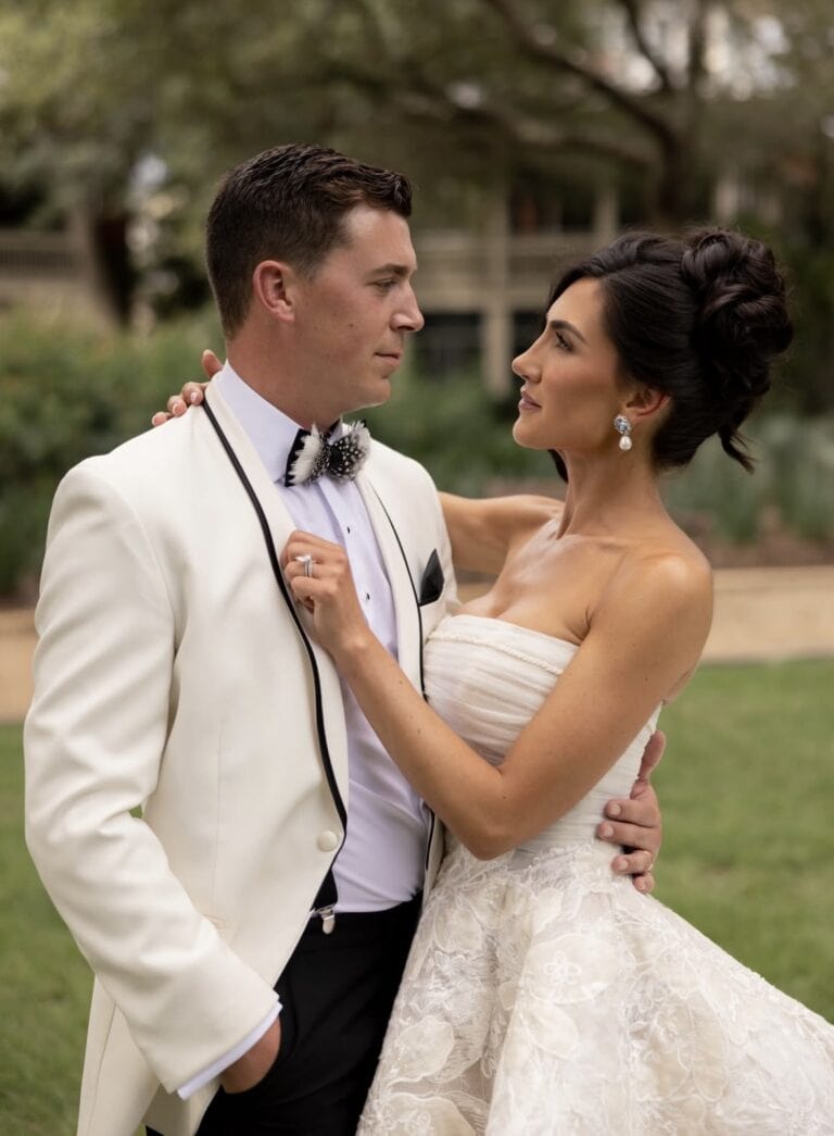 Screenshot | National Tuxedo Rentals Bride in a lace wedding gown and groom in a white tuxedo embrace outdoors at a garden wedding.