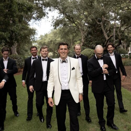 Groom in a white tuxedo jacket leads a group of groomsmen in black tuxedos across a grassy lawn.