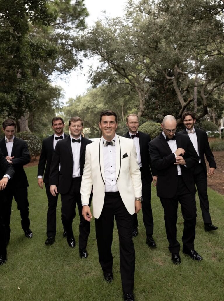 Screenshot | National Tuxedo Rentals Groom in a white tuxedo jacket leads a group of groomsmen in black tuxedos across a grassy lawn.