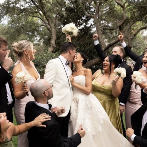 Bride and groom kiss while surrounded by bridesmaids and groomsmen holding white bouquets outdoors at a wedding reception stance.