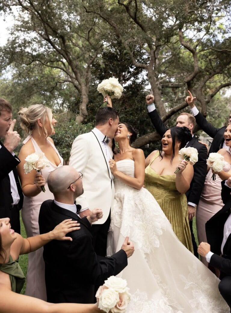 Screenshot | National Tuxedo Rentals Bride and groom kiss while surrounded by bridesmaids and groomsmen holding white bouquets outdoors at a wedding reception stance.